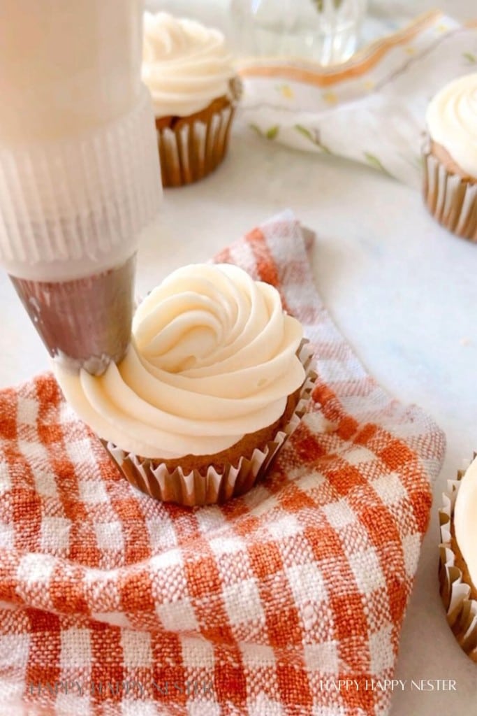 A close-up of a cupcake being frosted with a swirl of creamy white icing. The cupcake is placed on a red and white checkered cloth. Other frosted cupcakes are visible in the background.