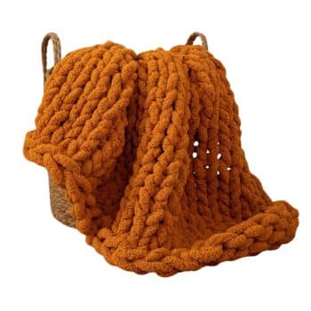 A large, chunky-knit orange blanket draped over the edge of a woven basket. The blanket's thick, bulky yarn is loosely knit, creating a cozy and textured appearance. The basket has two handles, suggesting it serves as both storage and decoration.