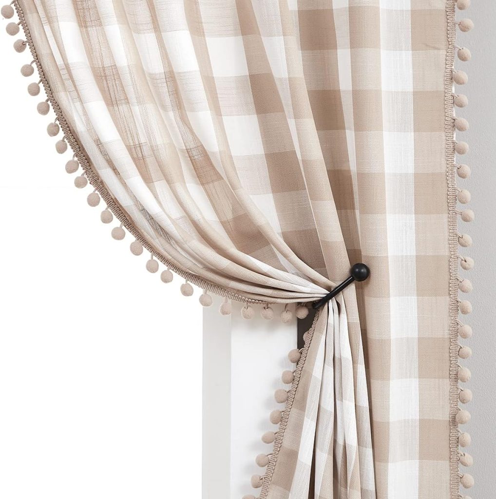 A light beige and white checkered curtain adorned with a fringe of small pom-poms is pulled to the side and held by a black curtain tieback on a white wall. The fabric has a soft, flowing texture and adds a touch of cozy, rustic charm to your festive fall decor.