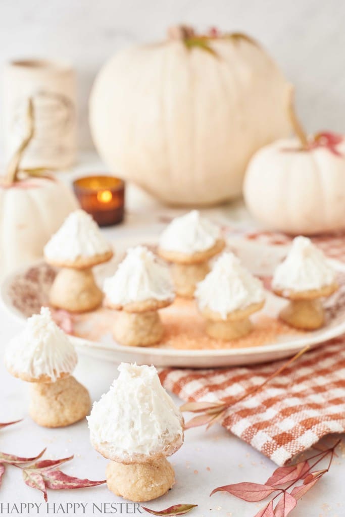 A decorative arrangement of mushroom-shaped autumn cookies with white frosting, displayed on a white plate. Around the plate, there are fall leaves, a plaid cloth, and white pumpkins, creating a cozy autumn atmosphere. A small lit candle is visible in the background.