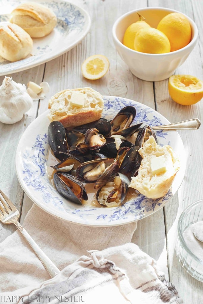 A white plate with a blue pattern features a dish of steamed mussels, served with slices of bread and pats of butter. On the table are a bowl of yellow lemons, a head of garlic, and additional bread rolls. A cut lemon and utensils are also present, reflecting bright natural light—the perfect setting for autumn recipes.