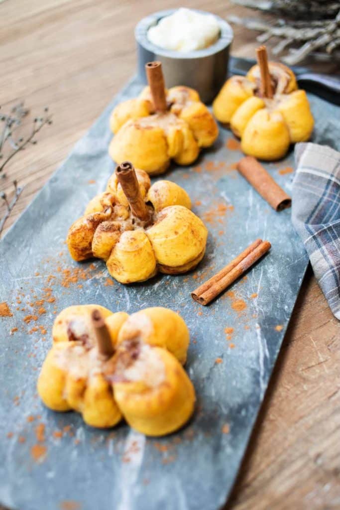 Four pumpkin-shaped, gluten-free pastries with cinnamon sticks in the center are displayed on a slate board. The pastries are dusted with cinnamon, and cinnamon sticks are placed nearby. A bowl of whipped cream is in the background, and a plaid cloth is partially visible.