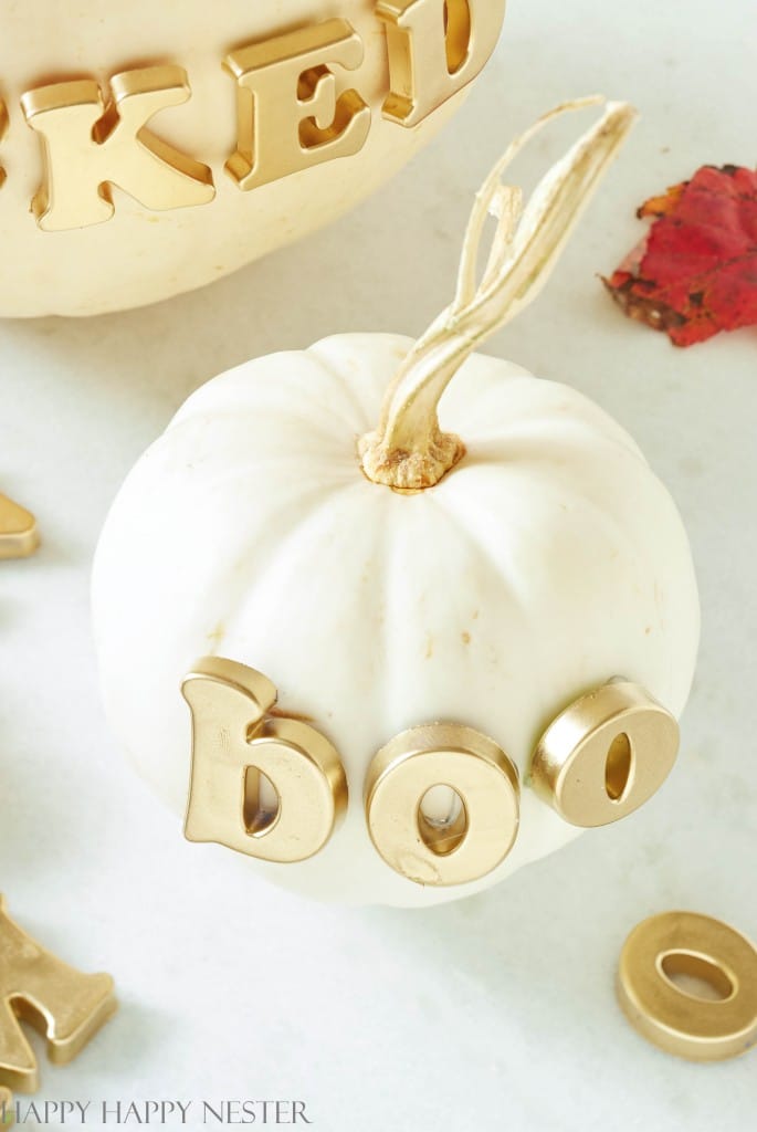 A small white pumpkin with gold letters spelling "BOO" on its front sits on a light surface. In the background, another larger pumpkin, also decorated with gold letters, is partially visible. A red autumn leaf lies nearby, adding to your fall decor ideas.