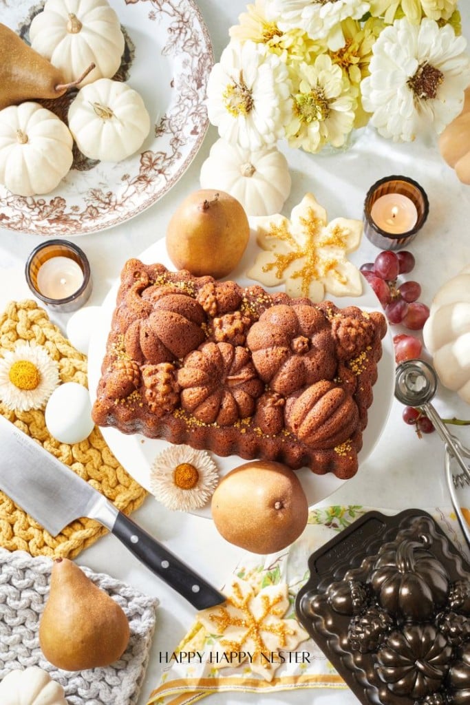 A beautifully decorated table with a fall-themed cake featuring pumpkin shapes, surrounded by pears, mini white pumpkins, flowers, a knife on a cutting board, and a crocheted mat. The scene is accentuated with autumn leaves and festive decor.