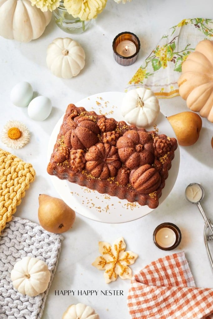 A fall-themed table setting features a pumpkin-shaped loaf cake on a white cake stand. Surrounding it are white pumpkins, pears, candles, eggs, and decorative autumn items such as knitted cloths and flowers. A checkered napkin and linen are also present.