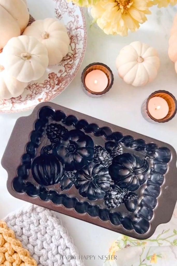 A fall-themed scene featuring a baking mold with intricate patterns of pumpkins and leaves. Surrounding the mold are white pumpkins, lit candles in brown holders, a yellow flower arrangement, and cozy knitted cloths in white and mustard yellow hues.