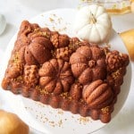 A richly decorated pumpkin-shaped bundt cake sits on a white plate, surrounded by white mini pumpkins, pears, and lit candles. Gold sprinkles are scattered on and around the cake. A patterned cloth and a candle in a glass holder are also visible.