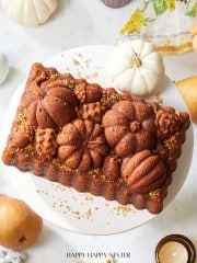 A richly decorated pumpkin-shaped bundt cake sits on a white plate, surrounded by white mini pumpkins, pears, and lit candles. Gold sprinkles are scattered on and around the cake. A patterned cloth and a candle in a glass holder are also visible.