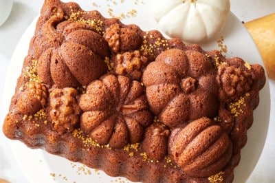 A richly decorated pumpkin-shaped bundt cake sits on a white plate, surrounded by white mini pumpkins, pears, and lit candles. Gold sprinkles are scattered on and around the cake. A patterned cloth and a candle in a glass holder are also visible.