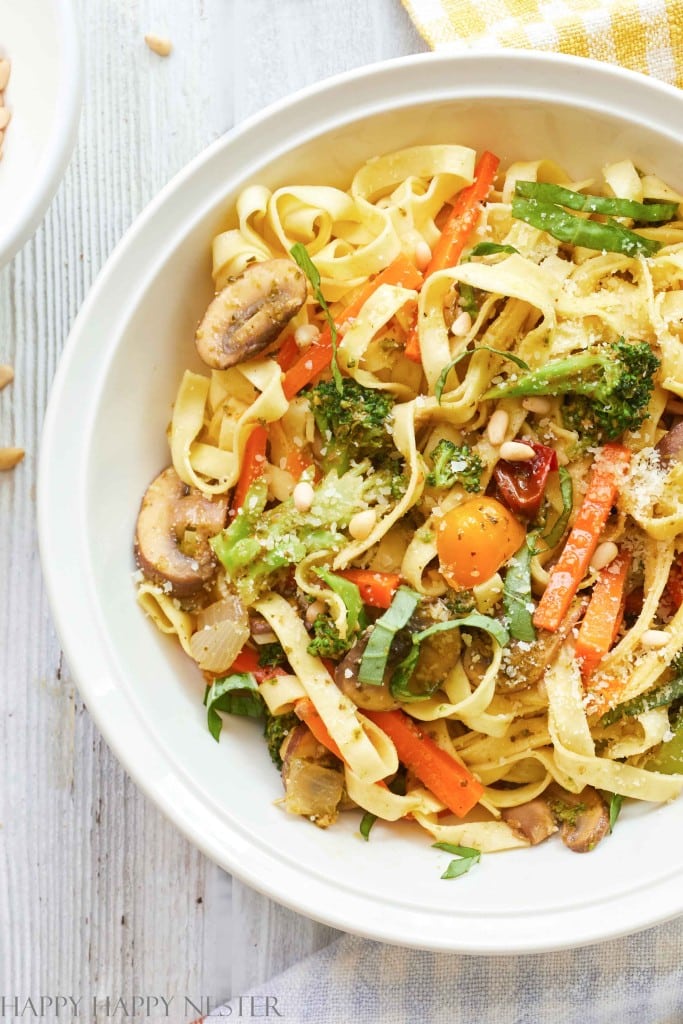 A bowl of colorful vegetable pasta featuring fettuccine, sliced carrots, broccoli florets, mushrooms, and cherry tomatoes, topped with grated cheese and garnished with fresh herbs. This perfect addition to your autumn recipes sits on a light wooden surface next to a yellow and white striped cloth.