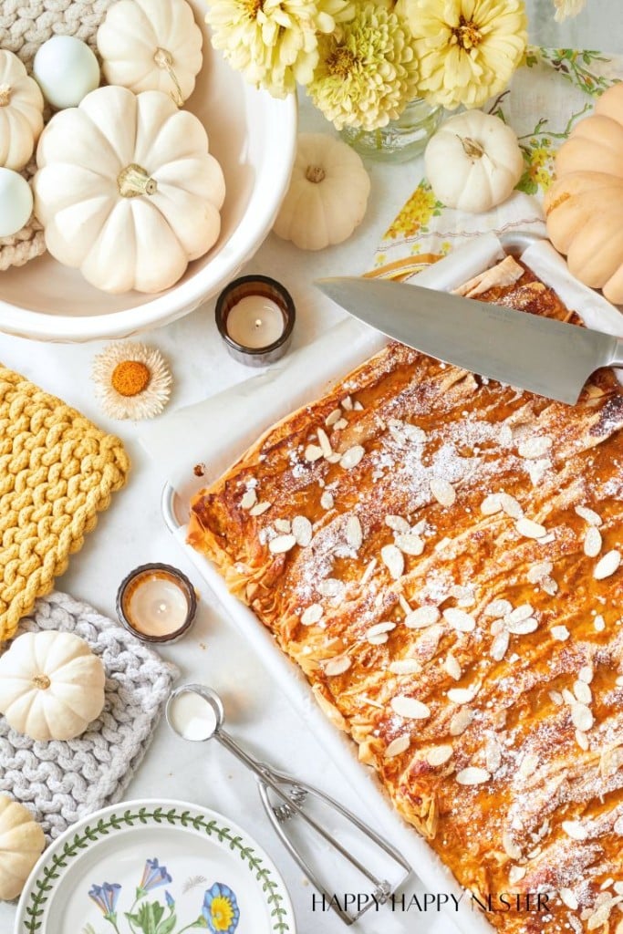 A top-view of a cozy autumn-themed table setting featuring a large freshly baked pastry topped with almonds and powdered sugar, surrounded by white pumpkins, candles, yellow and white flowers, and pastel crochet pieces. A knife rests near the pastry.