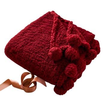 A folded red blanket adorned with pom-poms is shown against a white background. The blanket appears soft and cozy. A peach-colored ribbon is partially visible underneath the blanket, adding a touch of elegance.