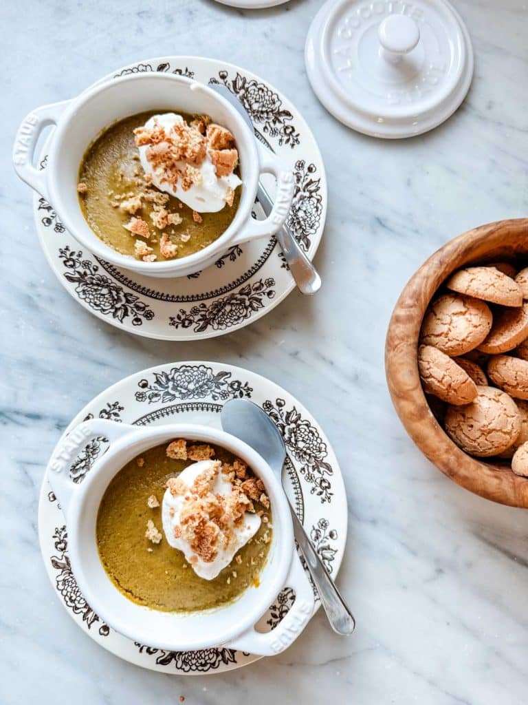 Two white ceramic bowls with ornate black patterns contain green soup topped with dollops of cream and crumbled cookies. A metal spoon rests on the rim of each bowl. Nearby, a wooden bowl brims with gluten-free cookies, while a white ceramic container with a lid adds a touch of elegance.