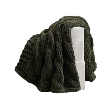 A chunky, dark green knitted blanket with a cable design is draped over a white block. The thick yarn and intricate pattern give the blanket a cozy and textured appearance.