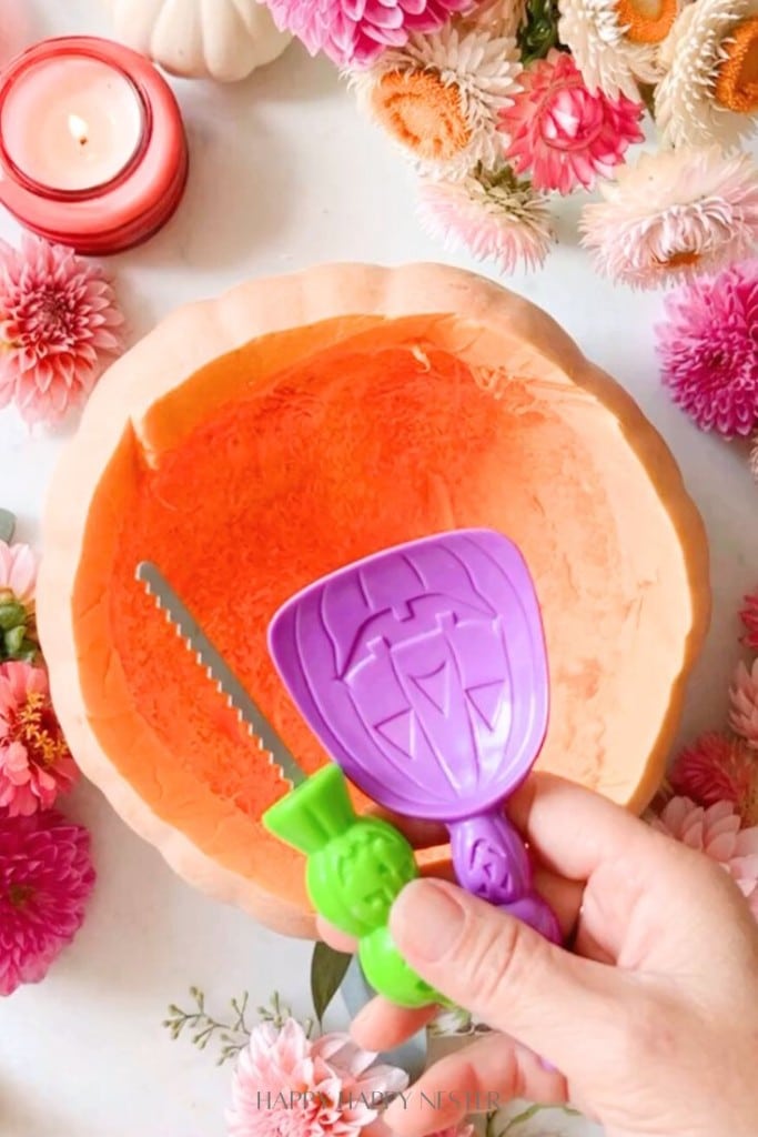 A hand holding a purple pumpkin carving scoop and a green serrated carving tool in front of an orange Halloween pumpkin bucket. The scene, perfect for a DIY project, is decorated with pink, orange, and white flowers. A red candle is visible nearby, completing this festive flower pumpkin centerpiece.