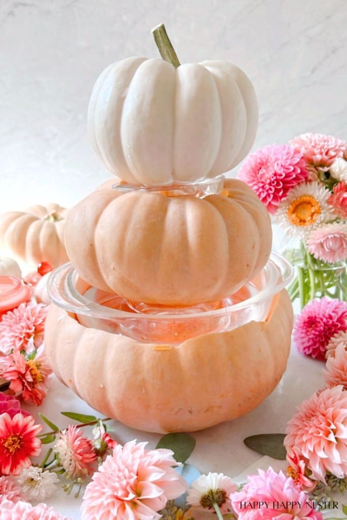 Two stacked pink pumpkins, with a white pumpkin on top, form a tiered decoration on a white surface, surrounded by various pink and white flowers. The background is subtly textured, adding a touch of elegance to the autumn-inspired arrangement.