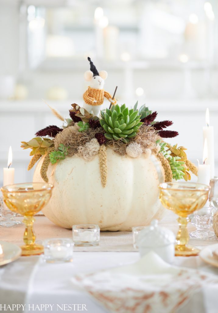 A centerpiece of a large white pumpkin decorated with green succulents, dried moss, and various dried plants is the epitome of fall decor ideas. The arrangement includes a small figurine at the top and is surrounded by lit candles and amber glasses on an elegantly set table.