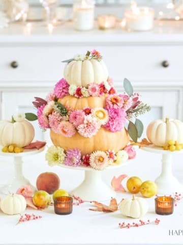 A festive table setting with a centerpiece featuring a tiered pumpkin arrangement adorned with pink, peach, and white flowers. Smaller pumpkins, peaches, and candles are placed around it. The background includes white cabinetry, candles, and fall-themed decor.