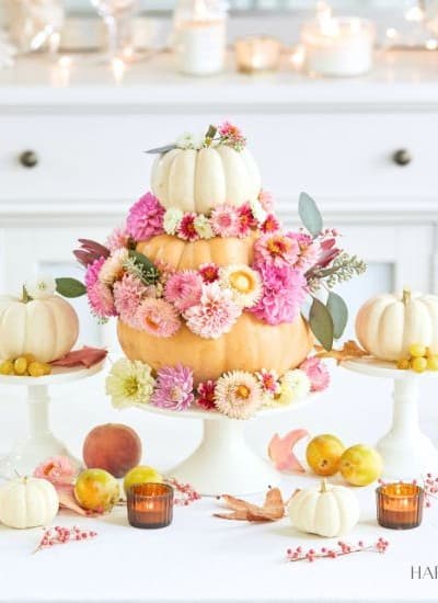 A festive table setting with a centerpiece featuring a tiered pumpkin arrangement adorned with pink, peach, and white flowers. Smaller pumpkins, peaches, and candles are placed around it. The background includes white cabinetry, candles, and fall-themed decor.