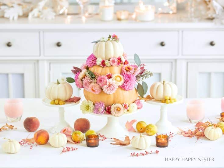 A festive table setting with a centerpiece featuring a tiered pumpkin arrangement adorned with pink, peach, and white flowers. Smaller pumpkins, peaches, and candles are placed around it. The background includes white cabinetry, candles, and fall-themed decor.