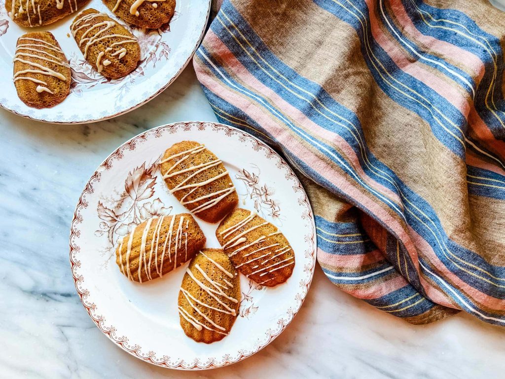 Three brown, oval-shaped fall cookies with white drizzle rest on a decorative plate, next to a blue and pink striped cloth. Another plate with similar cookies is partially visible above.
