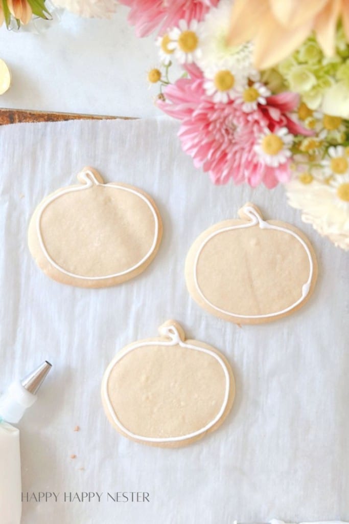 Three pumpkin-shaped sugar cookies on parchment paper, outlined with white icing. The cookies are surrounded by a variety of fresh flowers. A piping bag is seen in the corner of the image. The text "HAPPY HAPPY NESTER" is visible at the bottom left.