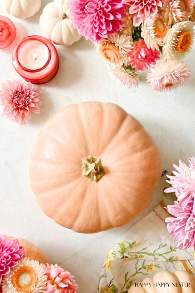 A light-colored pumpkin serves as the centerpiece, surrounded by pink and white flowers, a red candle, and a floral handkerchief. The overall DIY arrangement gives a cozy and decorative autumn feel.