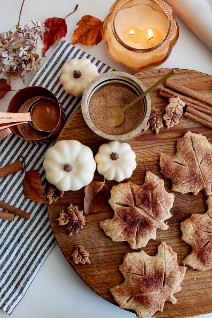 A wooden board displays leaf-shaped cookies alongside small white pumpkins and delicious gluten-free pumpkin cupcakes, a striped cloth with copper measuring spoons, loose spices, and cinnamon sticks. A lit candle in a glass jar and autumn leaves enhance the cozy seasonal atmosphere.