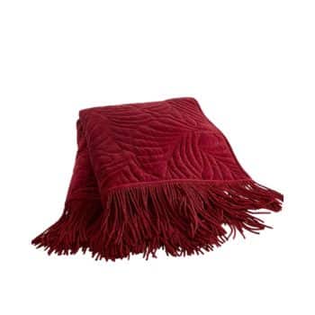 A folded, deep red quilted throw blanket with an intricate stitched pattern and fringed edges is neatly placed upright against a white background.