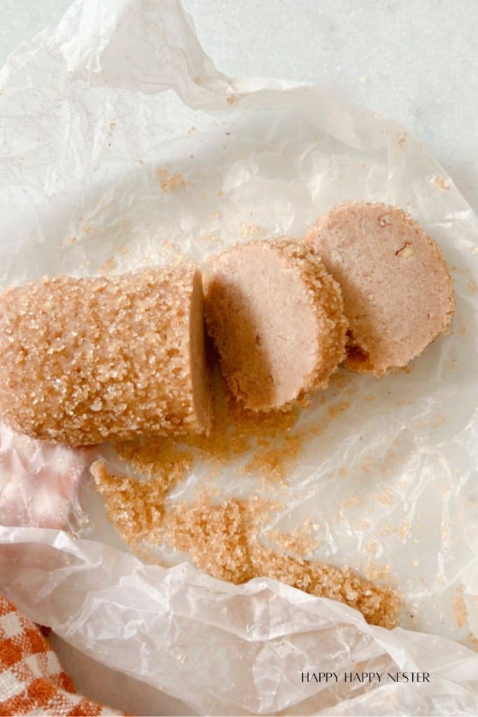 A cylindrical loaf of sliced brown sugar cookie dough rests on white parchment paper. Two slices are cut and laying flat, with sugar crystals scattered around. The background includes a red-and-white checkered cloth.