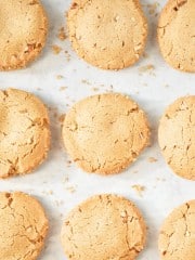 A close-up view of several evenly spaced, round, golden-brown cookies on a light surface, with a few crumbs scattered around. The cookies have a slightly cracked texture on top. The bottom right corner includes the text "HAPPY HAPPY NESTER.