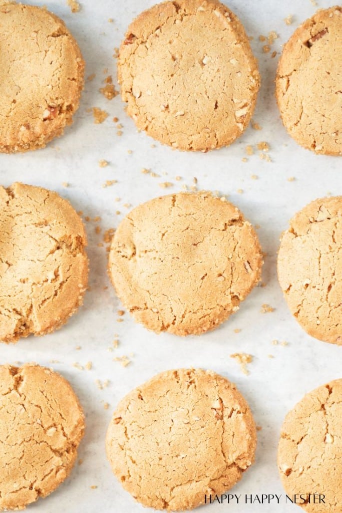 Eight almond flour autumn cookies with a golden-brown, slightly cracked surface are arranged in rows on a white parchment-lined baking sheet. Crumbs are scattered between the cookies. The phrase "HAPPY HAPPY NESTER" is printed in the bottom right corner.