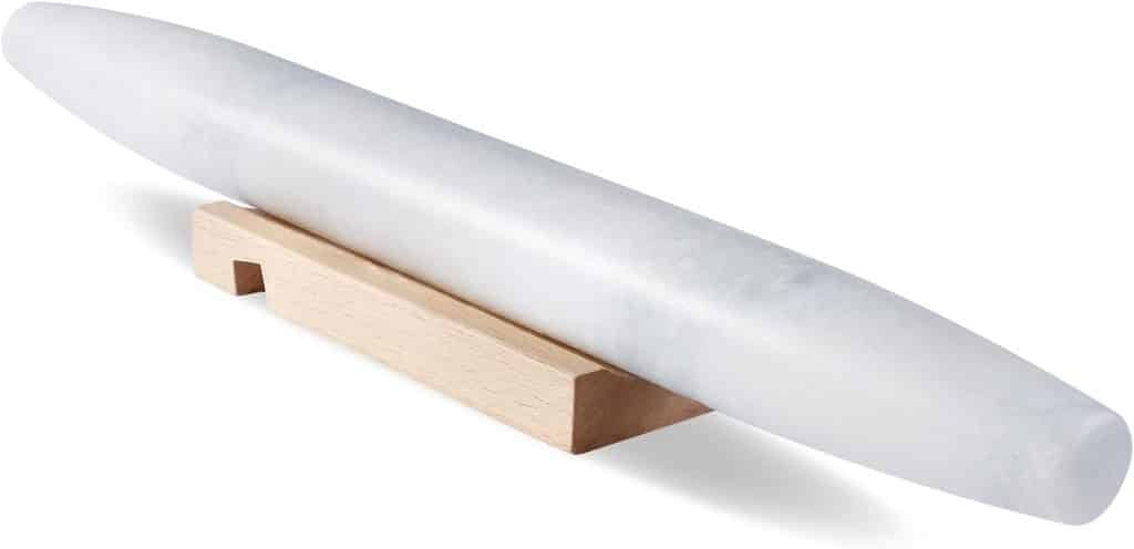 A white marble rolling pin with tapered ends, perfect for crafting fall cookies, rests on a wooden cradle, isolated on a white background.