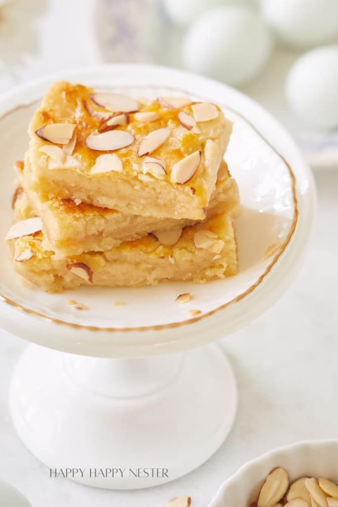 Two squares of almond-topped blondies sit stacked on a white pedestal plate. The blondies have a golden-brown top and are garnished with sliced almonds. The plate is set on a white surface with a soft focus background that includes blurred, pastel-colored decorations.