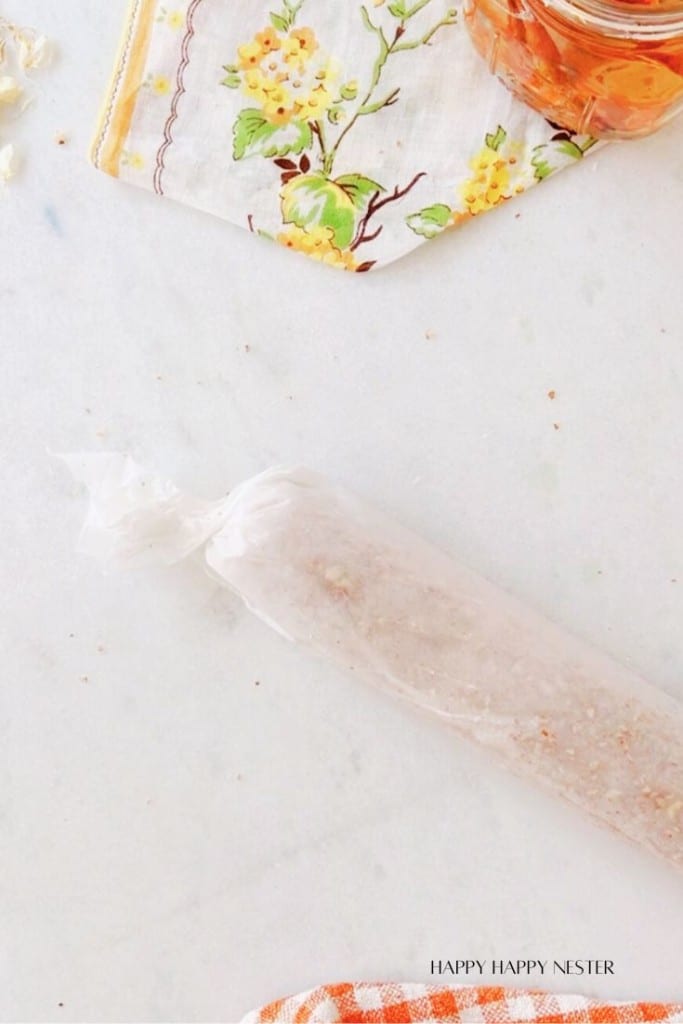 A cloth with floral patterns lies on a marble surface next to a jar with a golden liquid. A roll of food wrapped in plastic is placed on the marble surface, near the cloth. In the bottom-right corner, there is text that reads "Happy Happy Nester.