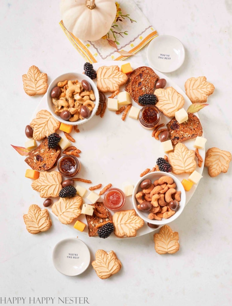 A festive charcuterie wreath arranged with various snacks, including cashews, berries, cheeses, cookies in autumn leaf shapes, and dips in small bowls on a white background. Decorated with a small pumpkin and a napkin in the center, this delightful creation is perfect for your autumn recipes collection.