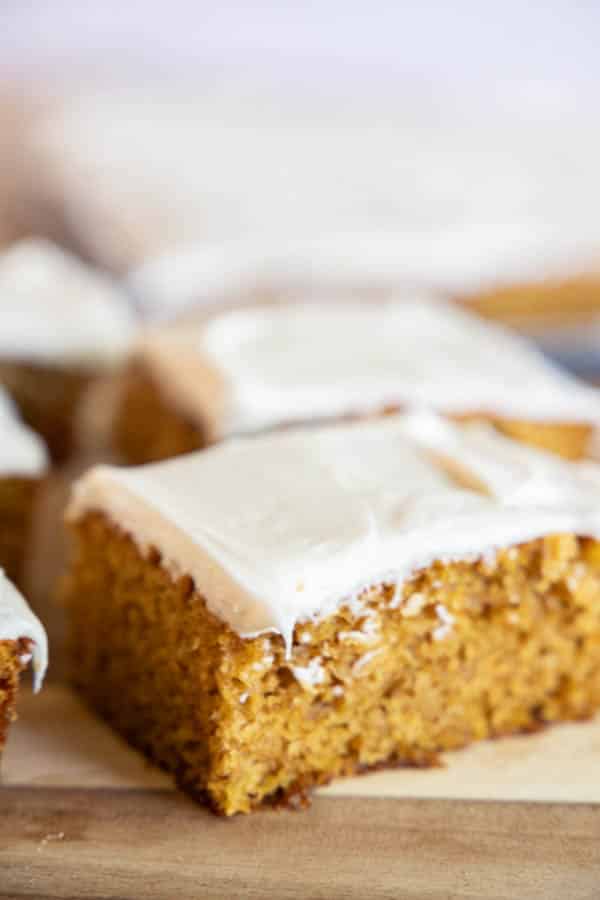 Close-up of a slice of moist pumpkin cake topped with a generous layer of creamy white icing. The slice reveals a fluffy, orange-brown interior that contrasts with the smooth, white frosting. The background shows more slices of the gluten-free pumpkin cake on a wooden surface.