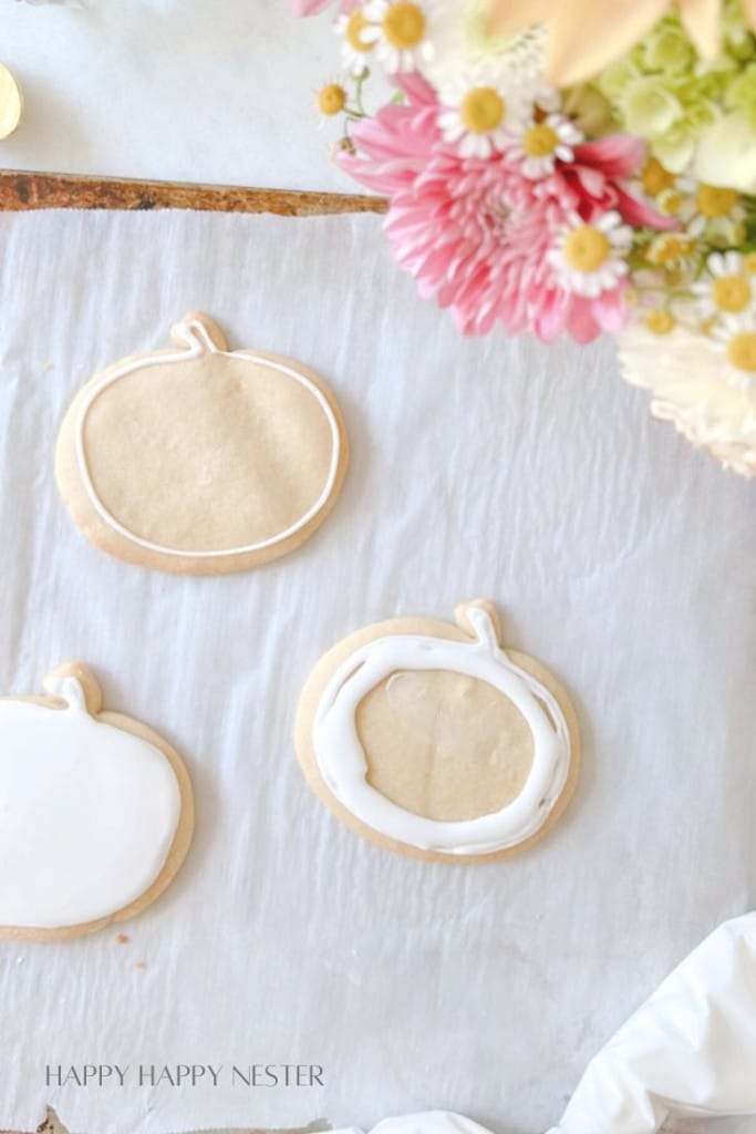 Three pumpkin-shaped cookies in various stages of being iced are placed on parchment paper. To the right is a vibrant bouquet of flowers with pink and white blooms. The text "HAPPY HAPPY NESTER" is visible at the bottom left corner of the image.