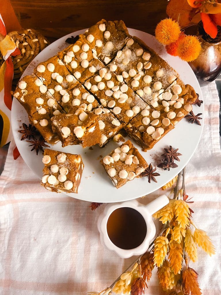 A plate of gluten free pumpkin blondies topped with white chocolate chips is sliced into squares and displayed on a white cloth with autumn decorations, including sprigs of wheat and orange flowers. A white mug filled with a warm brown beverage is placed nearby.