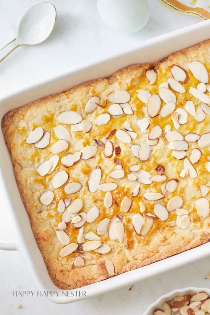 A rectangular white baking dish filled with a golden-brown almond cake topped with sliced almonds. The cake has a slightly textured surface with visible almond slices. Two spoons and part of a white plate edge can be seen nearby.