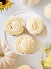 Three vanilla cupcakes with swirled frosting are placed on a round white stand. The surrounding area has white pumpkins and yellow flowers, giving a festive and bright appearance.