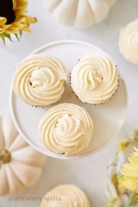 Three vanilla cupcakes with swirled frosting are placed on a round white stand. The surrounding area has white pumpkins and yellow flowers, giving a festive and bright appearance.