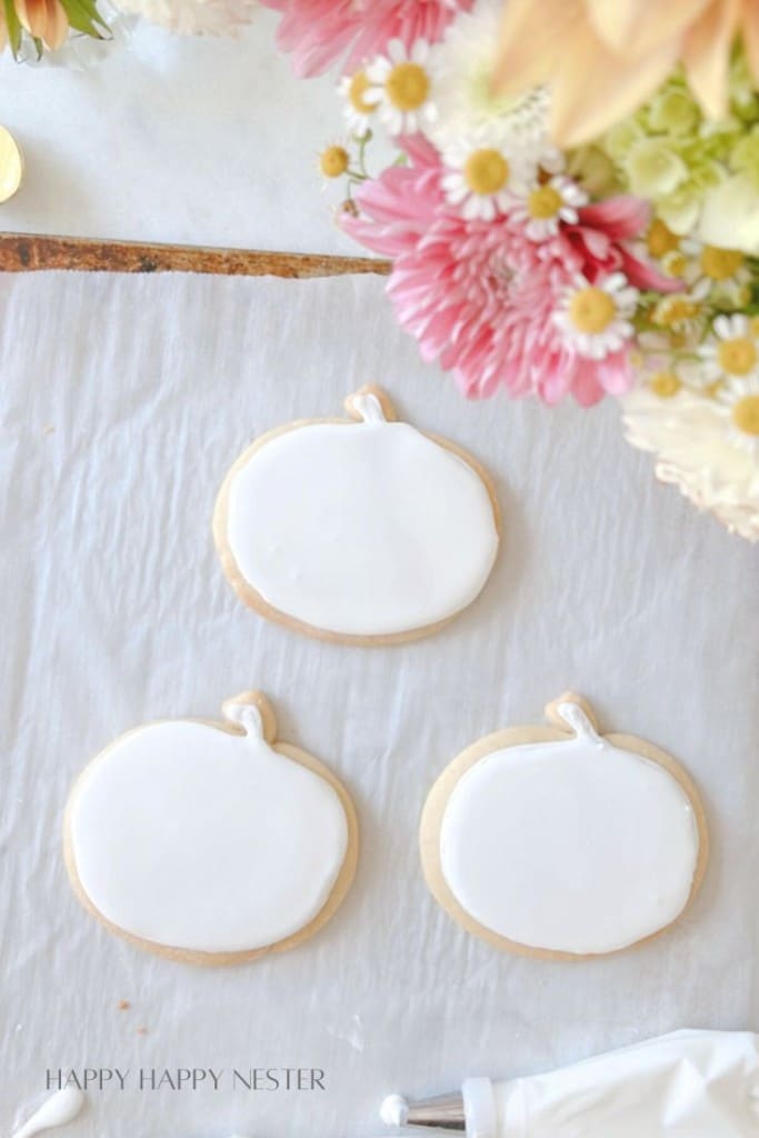 Three pumpkin-shaped cookies with white icing are placed on a sheet of parchment paper. A bouquet of colorful flowers partially frames the top right corner. The phrase "Happy Happy Nester" is lightly visible in the lower left corner.