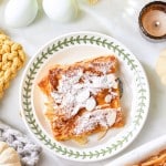 A plate with powdered sugar-dusted flaky pastry, surrounded by candles, white decorative pumpkins, eggs, and yellow flowers on a white surface. Cozy knitted items are also present, adding to the warm ambiance.