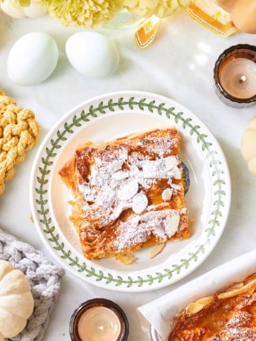 A plate with powdered sugar-dusted flaky pastry, surrounded by candles, white decorative pumpkins, eggs, and yellow flowers on a white surface. Cozy knitted items are also present, adding to the warm ambiance.