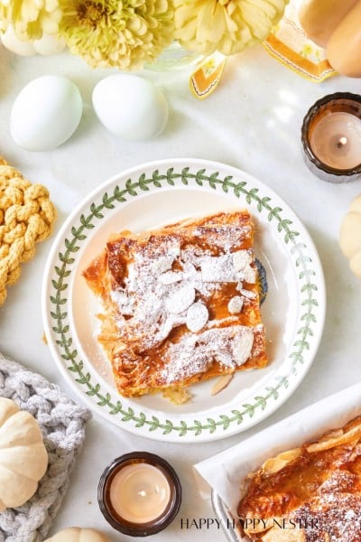 A plate with powdered sugar-dusted flaky pastry, surrounded by candles, white decorative pumpkins, eggs, and yellow flowers on a white surface. Cozy knitted items are also present, adding to the warm ambiance.