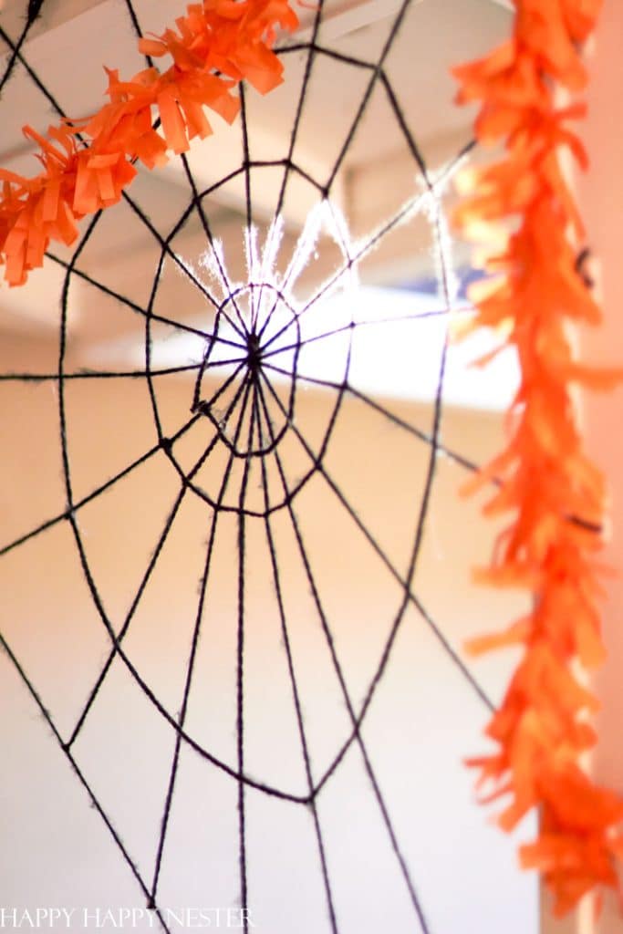 A homemade Halloween decoration featuring a black string spider web hanging from the ceiling, bordered with bright orange paper streamers. The web is intricately woven, and the background is softly blurred, making it a perfect piece of festive fall decor.