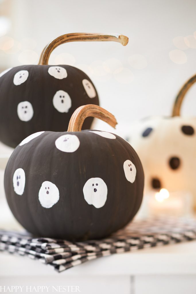 Two pumpkins are painted black and decorated with small white ghost faces. They are sitting on a black and white plaid cloth with blurred lights in the background, adding a festive Halloween touch to the festive fall decor. A third white pumpkin with black spots is partially visible.