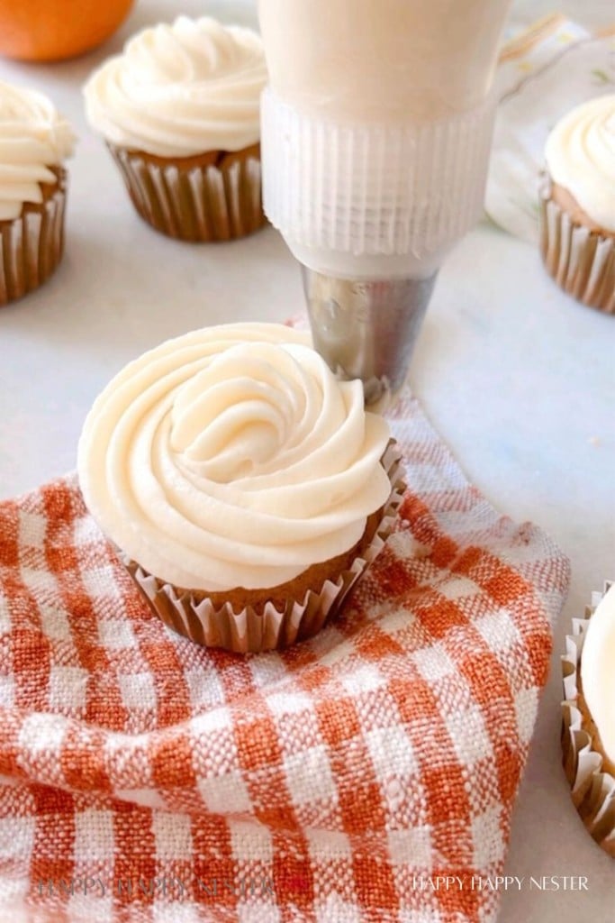 A close-up of a freshly frosted cupcake being decorated with white frosting from a piping bag. The cupcake sits on an orange and white checkered cloth, and there are several other frosted cupcakes visible in the background.