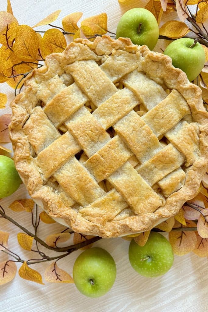 A lattice-topped apple pie, inspired by the cozy comfort of a Costco Pumpkin Pie Recipe, is surrounded by green apples and yellow autumn leaves on a light wooden surface. The golden flaky crust perfectly complements the rustic fall theme.